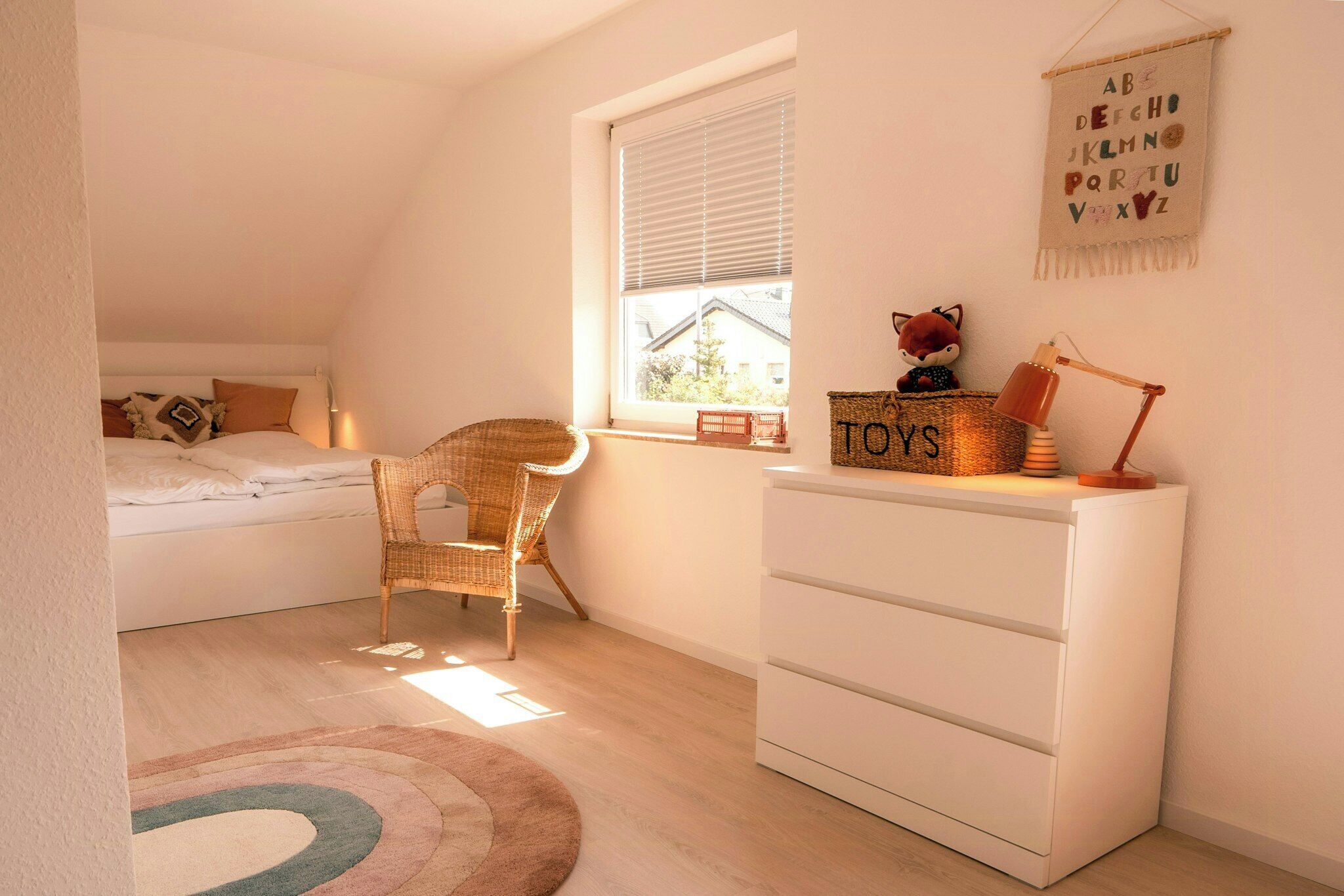 A child's bedroom with a bed, a wicker chair, a white dresser, a window with blinds, and a wall with alphabet letters.