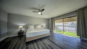 Ein Hotelzimmer mit einem großen Bett, einem Deckenventilator, einem Nachttisch mit Lampe und Blick auf ein Sportfeld durch eine Glastür.