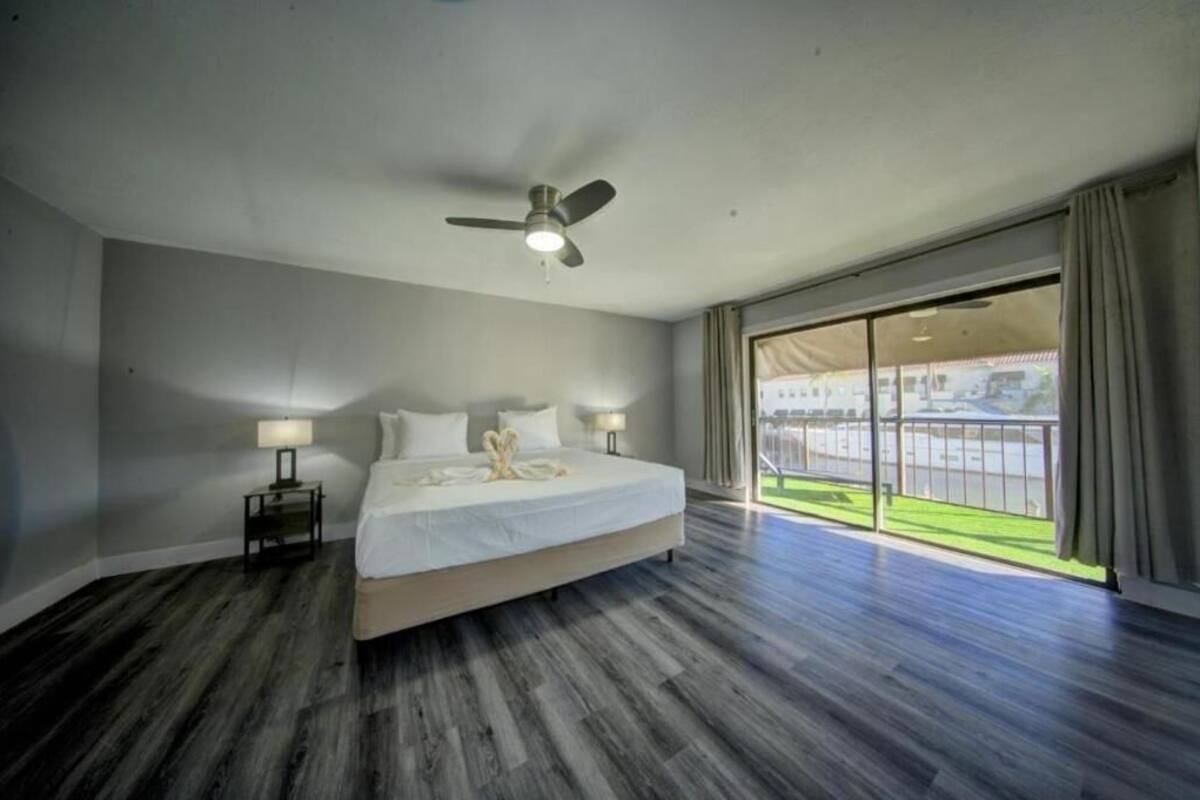 Quarto de hotel com uma cama grande, um ventilador de teto, uma mesa de cabeceira com abajur e vista para um campo esportivo através de uma porta de vidro deslizante.