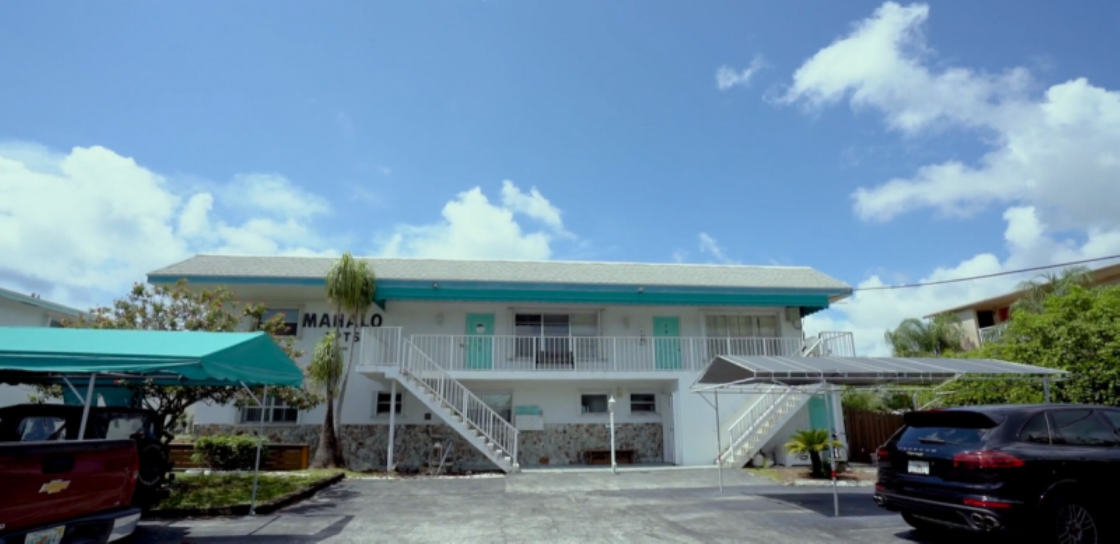 A two-story building with a staircase leading to the entrance, a covered parking area, and a clear sky.