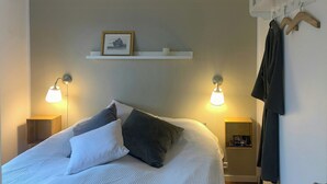 En snyggt bäddad säng med kuddar, två sänglampor, en inramad tavla på en hylla och en klädställning med en badrock.