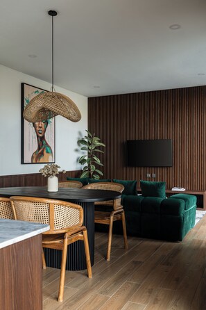 Un salon moderne comprenant un coin repas, un lambris en bois, un téléviseur à écran plat, des fauteuils en osier, un canapé vert et un luminaire suspendu.