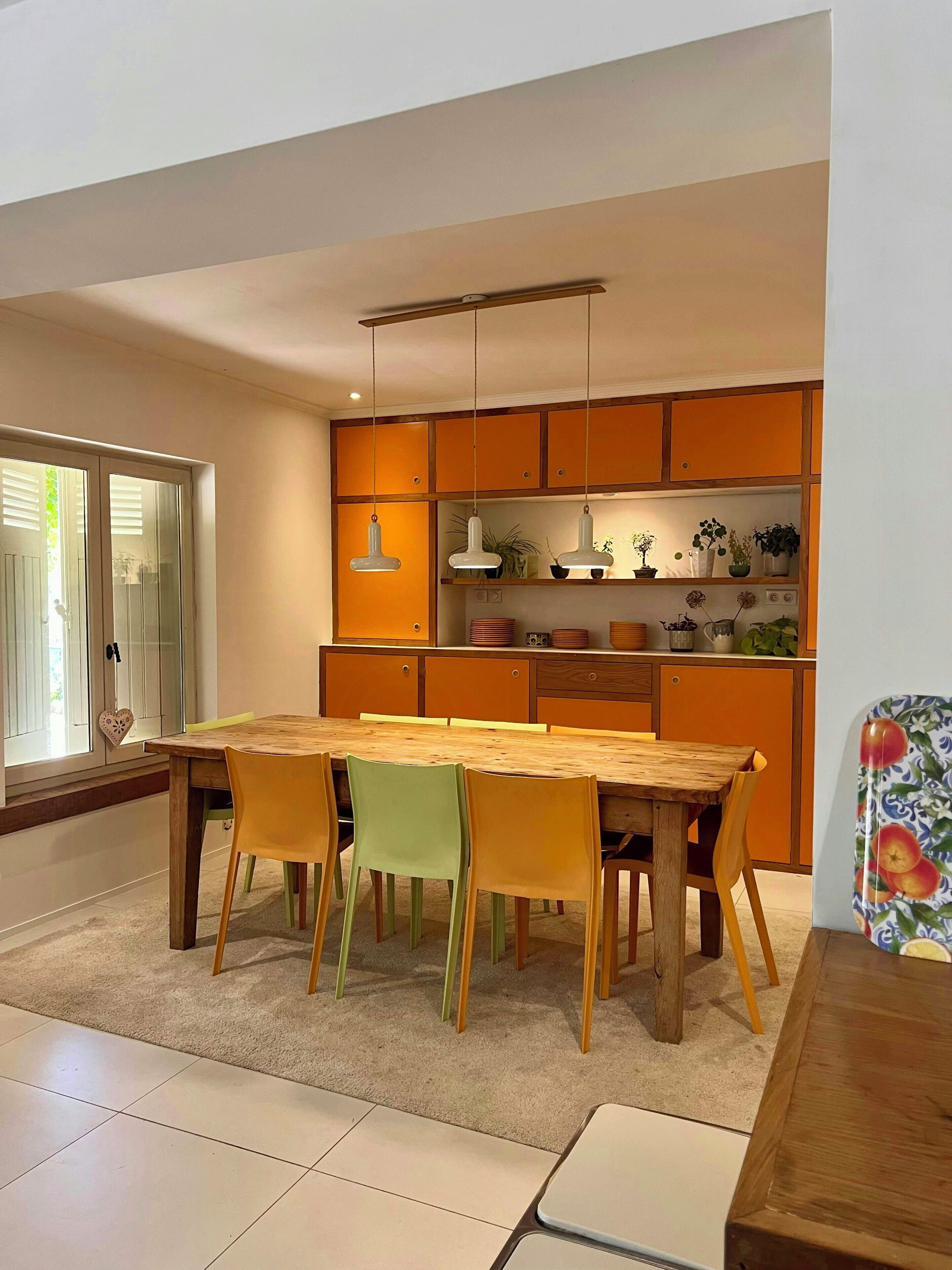 Uma sala de jantar com mesa e cadeiras de madeira, uma cozinha com armários laranja e uma janela com persianas.