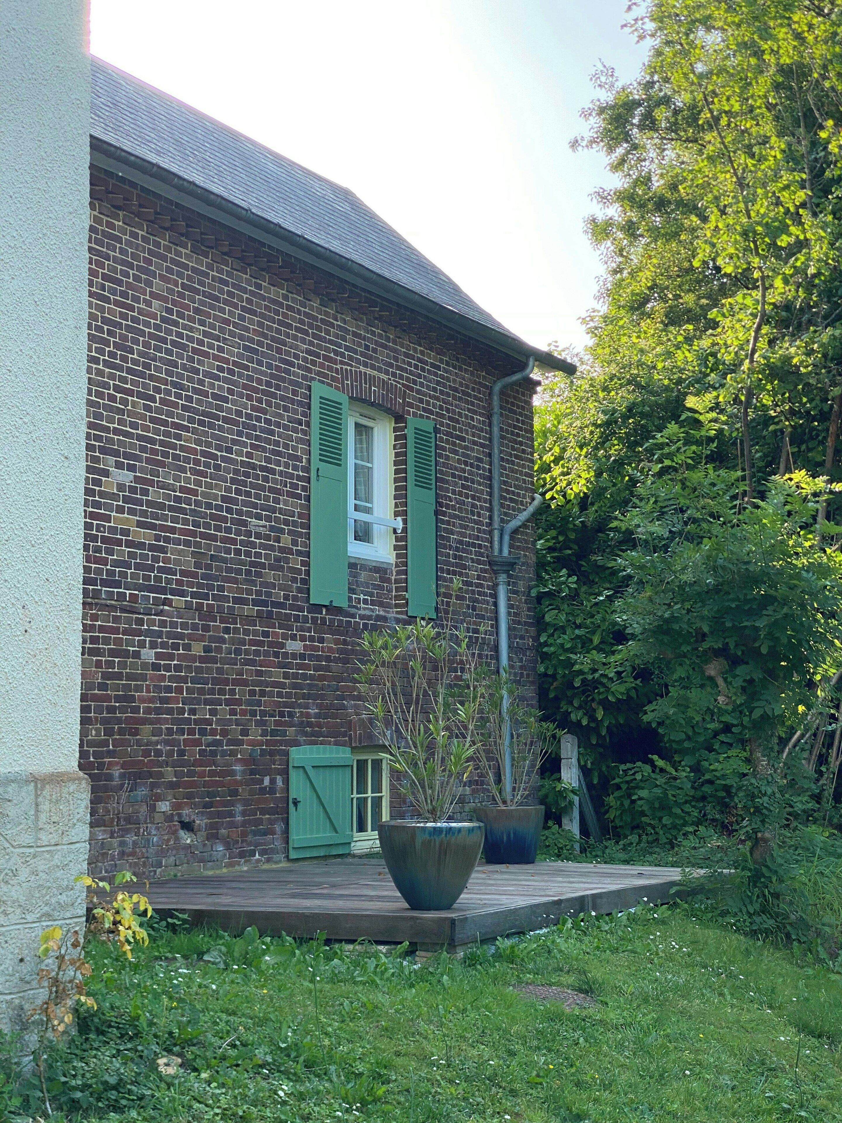 A brick building with a window and shutters, surrounded by greenery.