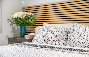 Ein Schlafzimmer mit einem Lattenrost, einem Bett mit weißer und schwarzer Punkte-Bettwäsche und einer Vase mit weißen Blumen auf einem Nachttisch.