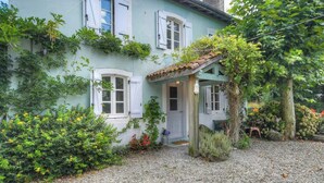 A light blue house with white shutters, a gravel driveway, and a garden with various plants and flowers. - Riverside house with farm animals in Béarn (Autevielle-Saint-Martin-Bideren)