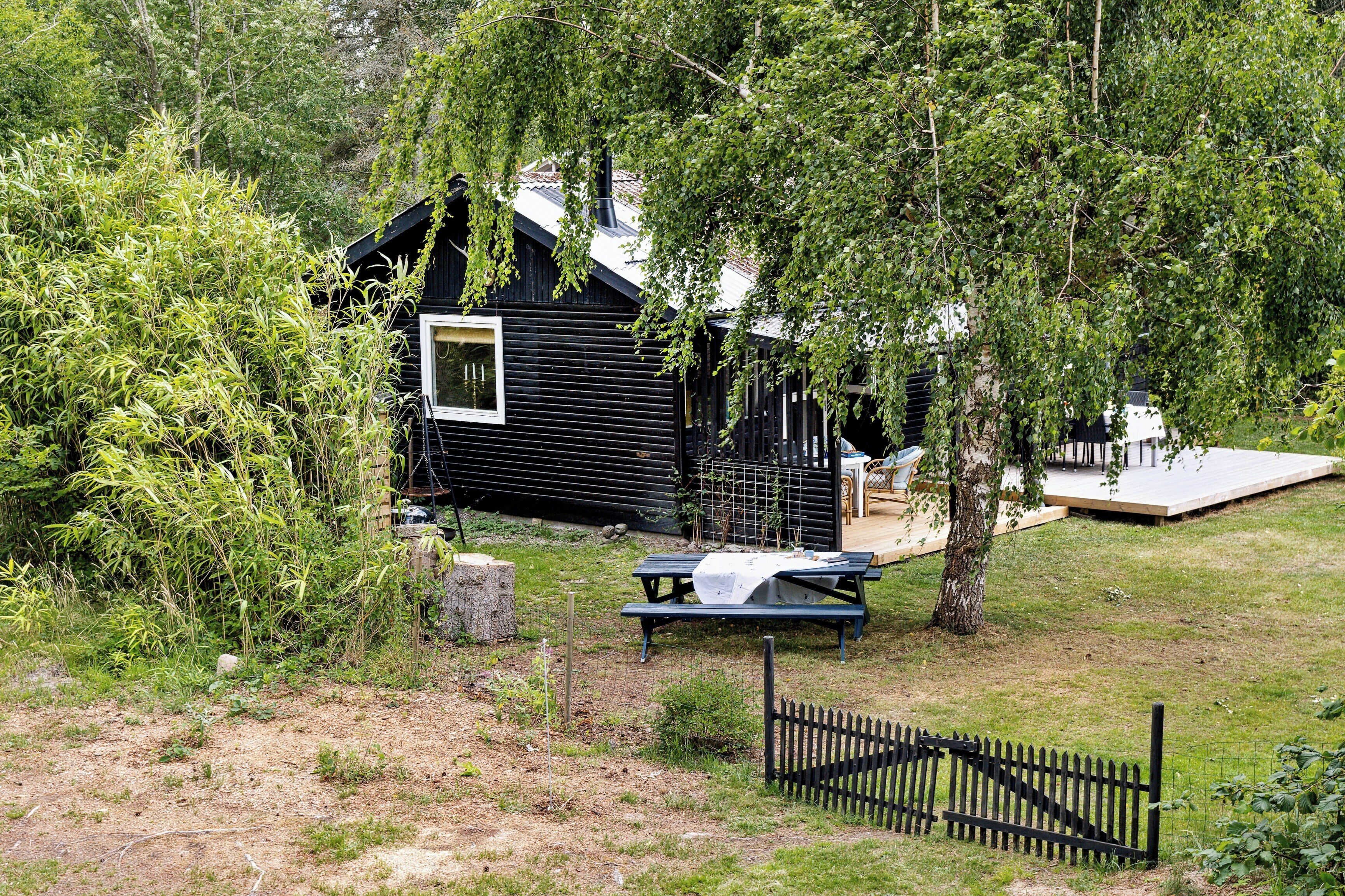 A black cabin with a picnic table and a fenced area, surrounded by greenery.