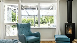 Een kamer met een blauwe fauteuil, een passende voetenbankje, een zwarte houtkachel en een groot raam met uitzicht op groen.
