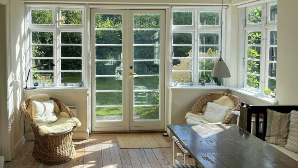 A sunlit room with a large sliding glass door, wicker furniture, a wooden table, and a view of greenery outside. - Villa from 1917 high above Tisvildeleje (Tisvilde)