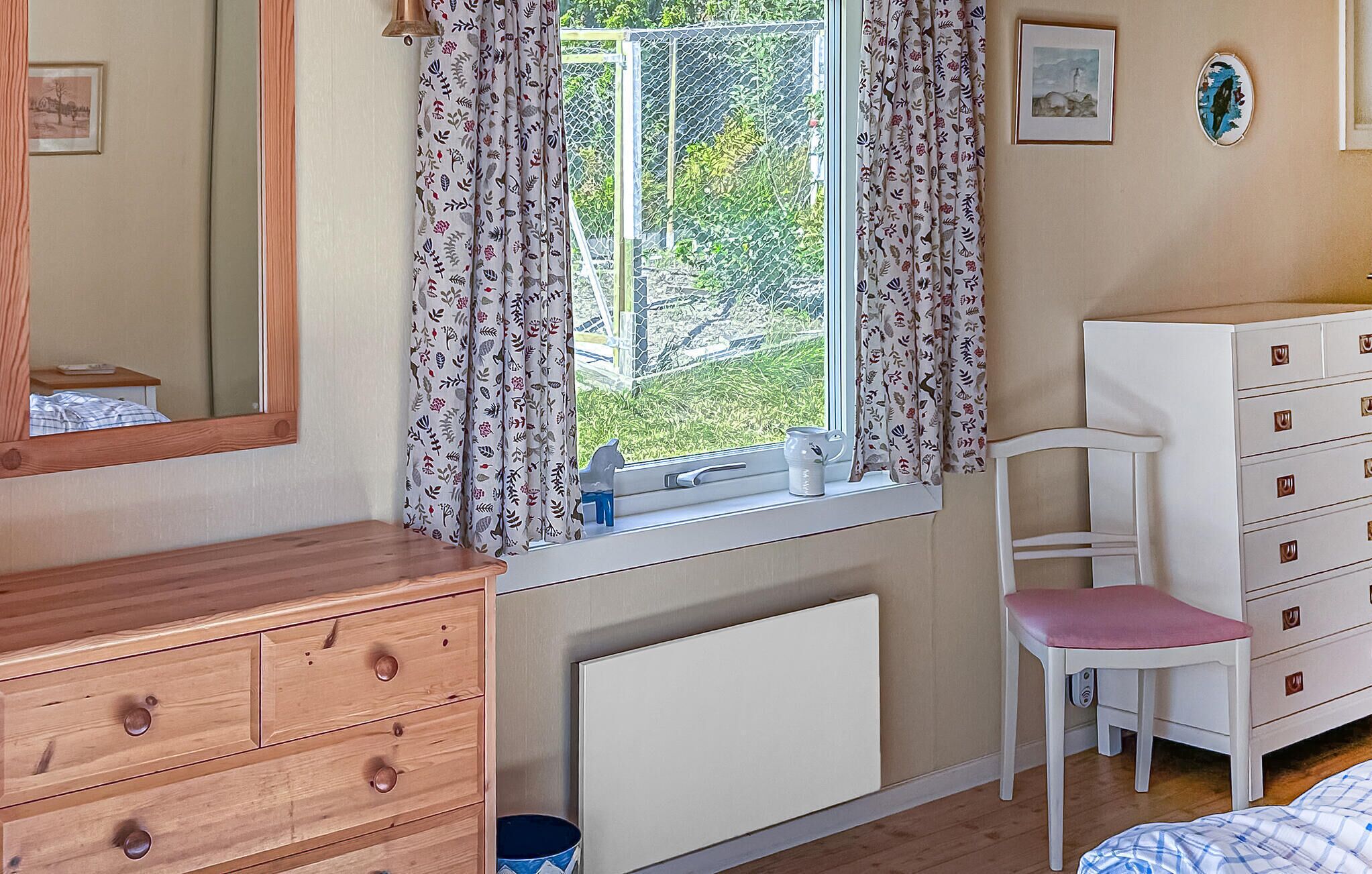 A bedroom with a wooden dresser, a white radiator, a window with floral curtains, a white chair, and a dresser with drawers.