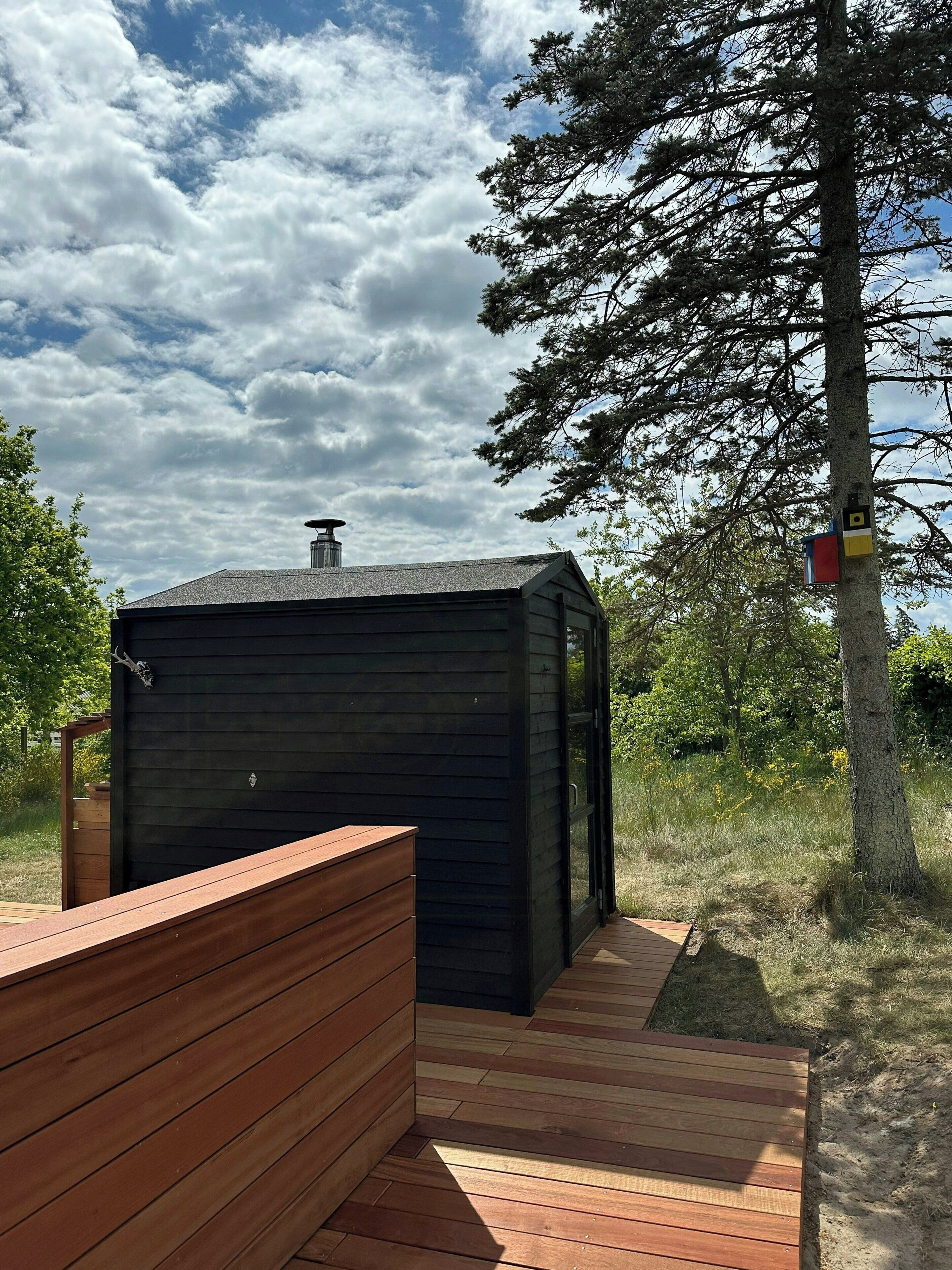 A small, dark wooden cabin with a chimney, a wooden deck, and a tree in the background.
