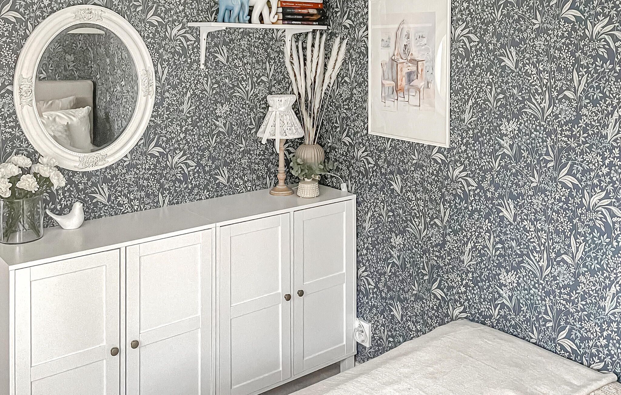 A bathroom with a white vanity, a mirror, a shelf with books, a vase with flowers, and a wall with floral wallpaper.