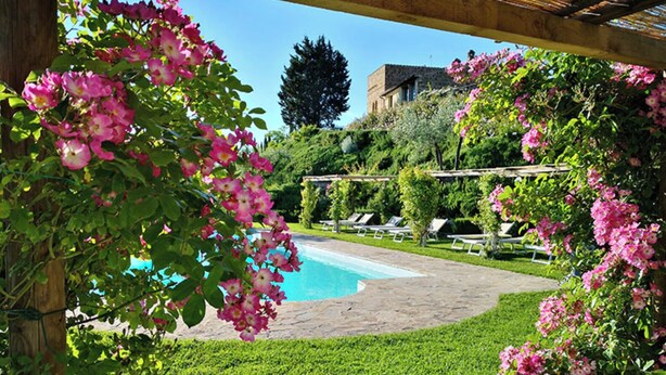 Zona de piscina amb una pèrgola, vegetació exuberante i un edifici al fons.