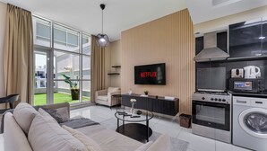 En moderne stue med sofa, sofabord, fjernsyn, køkkenområde med hårde hvidevarer og et stort vindue.