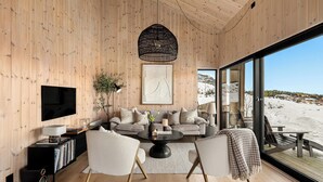 Ein modernes Wohnzimmer mit Holzvertäfelung, einem großen Fenster mit Blick auf eine verschneite Landschaft, einem Fernseher und einer gemütlichen Sitzecke.