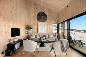 A modern living room with a wooden interior, a large window overlooking a snowy landscape, a television, and a cozy seating area. - Cabin overlooking Ryfylke fjord (Høyvik)