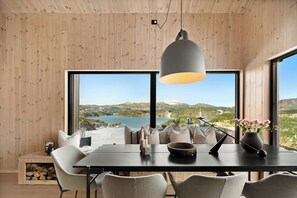 A dining area with a wooden table, chairs, a hanging light fixture, and a view of a lake and mountains through large windows. - Cabin overlooking Ryfylke fjord (Høyvik)