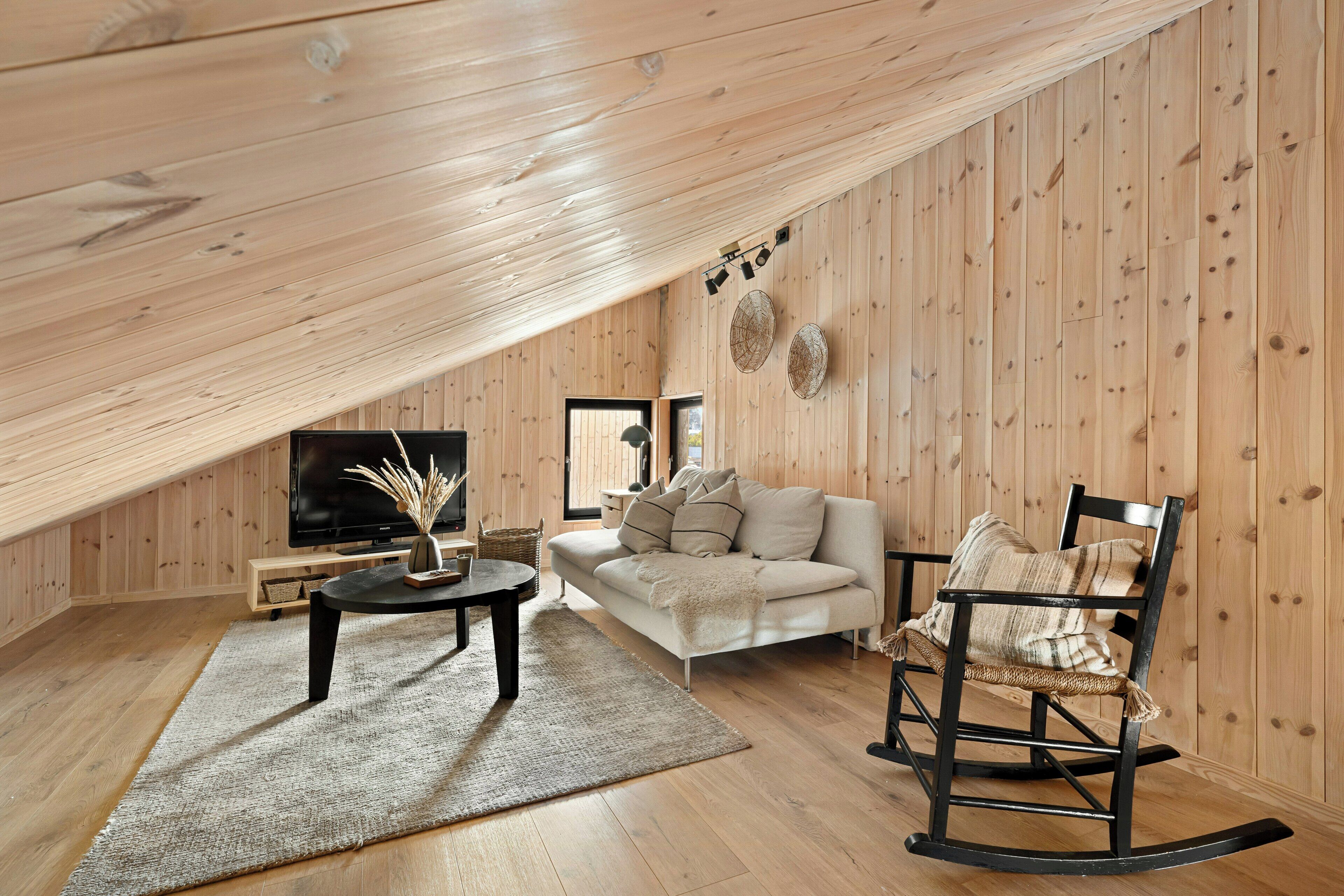 Ett rum i loftstil med ett träpanelat tak, en soffa, en gungstol, en tv och ett soffbord.