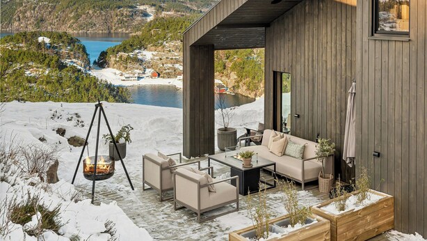 Ein moderner Außenbereich mit Holzterrasse, einer Sitzgelegenheit und Blick auf einen See und Berge.