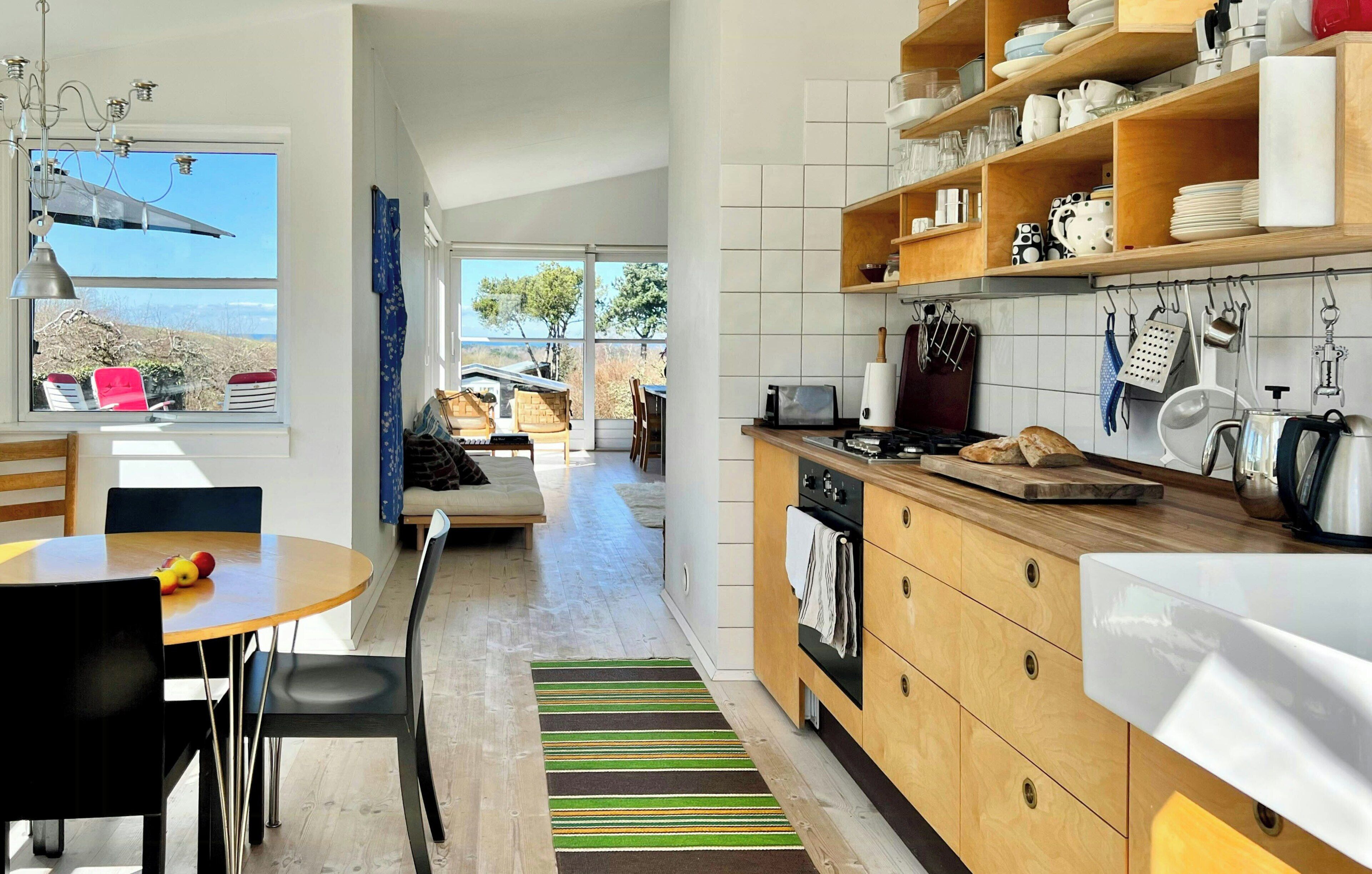 A modern kitchen with wooden cabinets, a dining area with a table and chairs, and a view through a large window.