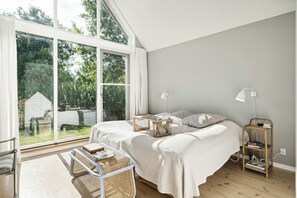 A bedroom with a large window, a bed with white bedding, a bedside table, a lamp, and a wicker chair. - Big family dream of a summer home (Ebeltoft)