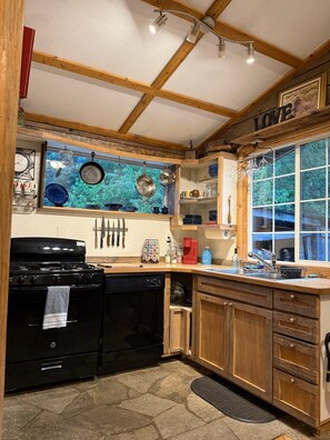 A kitchen with a stove, oven, and various utensils. There is a window with a view of trees, and wooden beams on the ceiling.