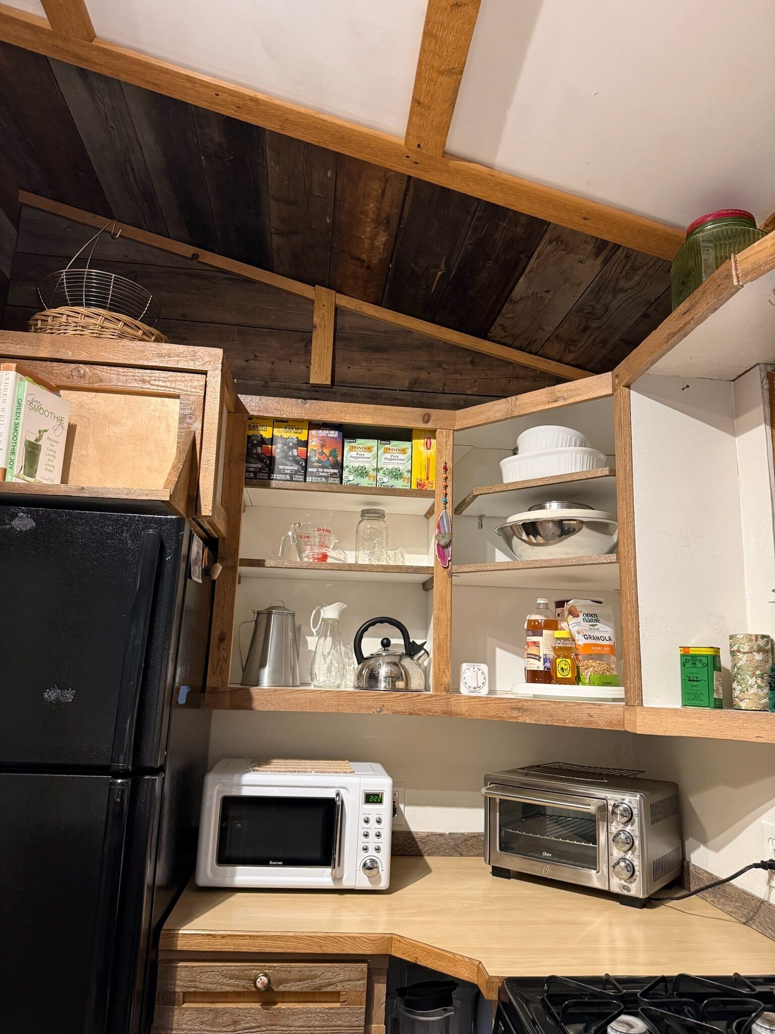 Una cucina compatta con un frigorifero nero, un forno a microonde, un tostapane ed scaffali in legno con diversi alimenti.