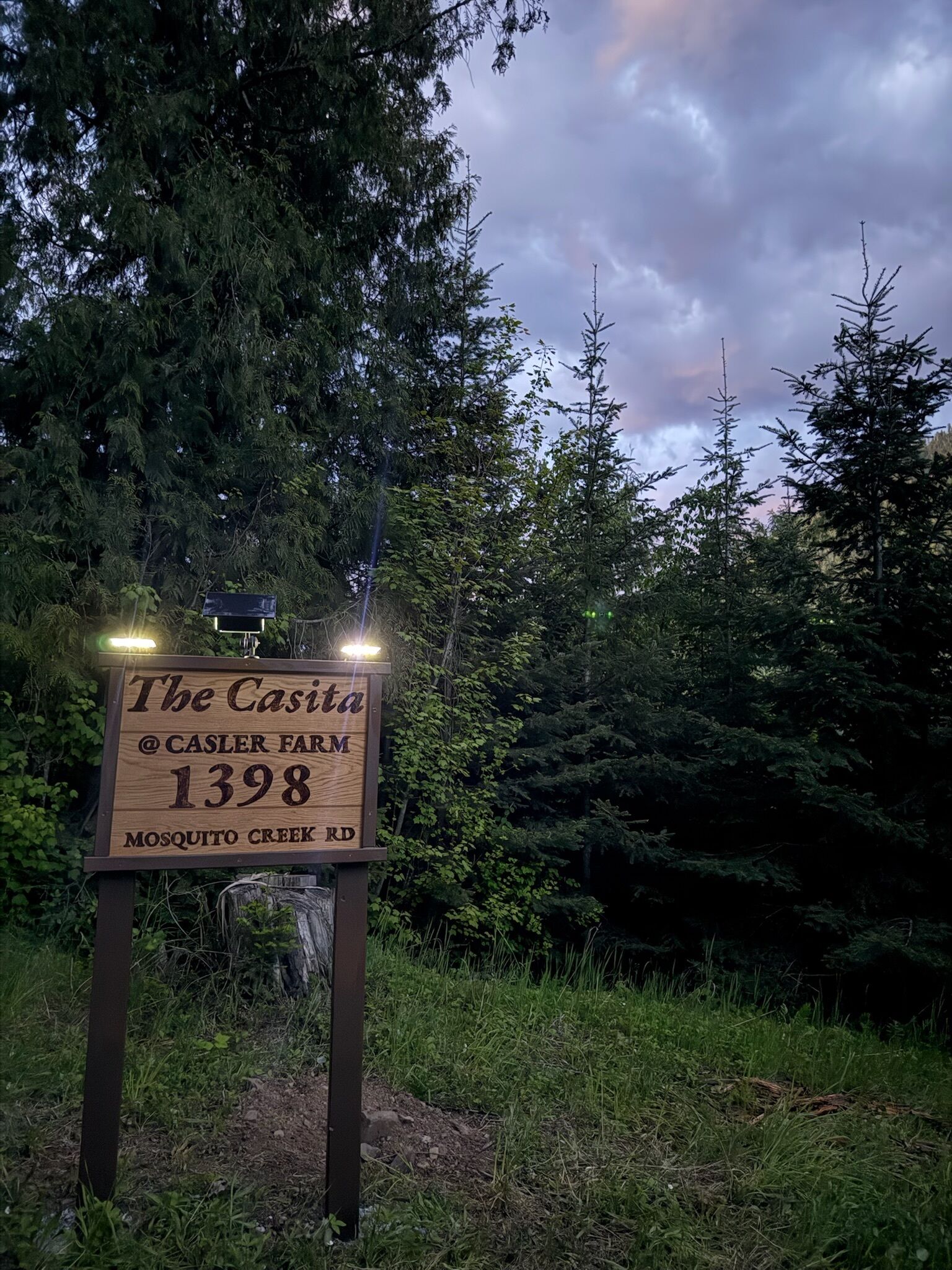 Ein hölzernes Schild mit der Aufschrift 'The Casa @ CASLER FARM 1398 MOSQUITO CREEK RD' vor Bäumen und einem bewölkten Himmel.