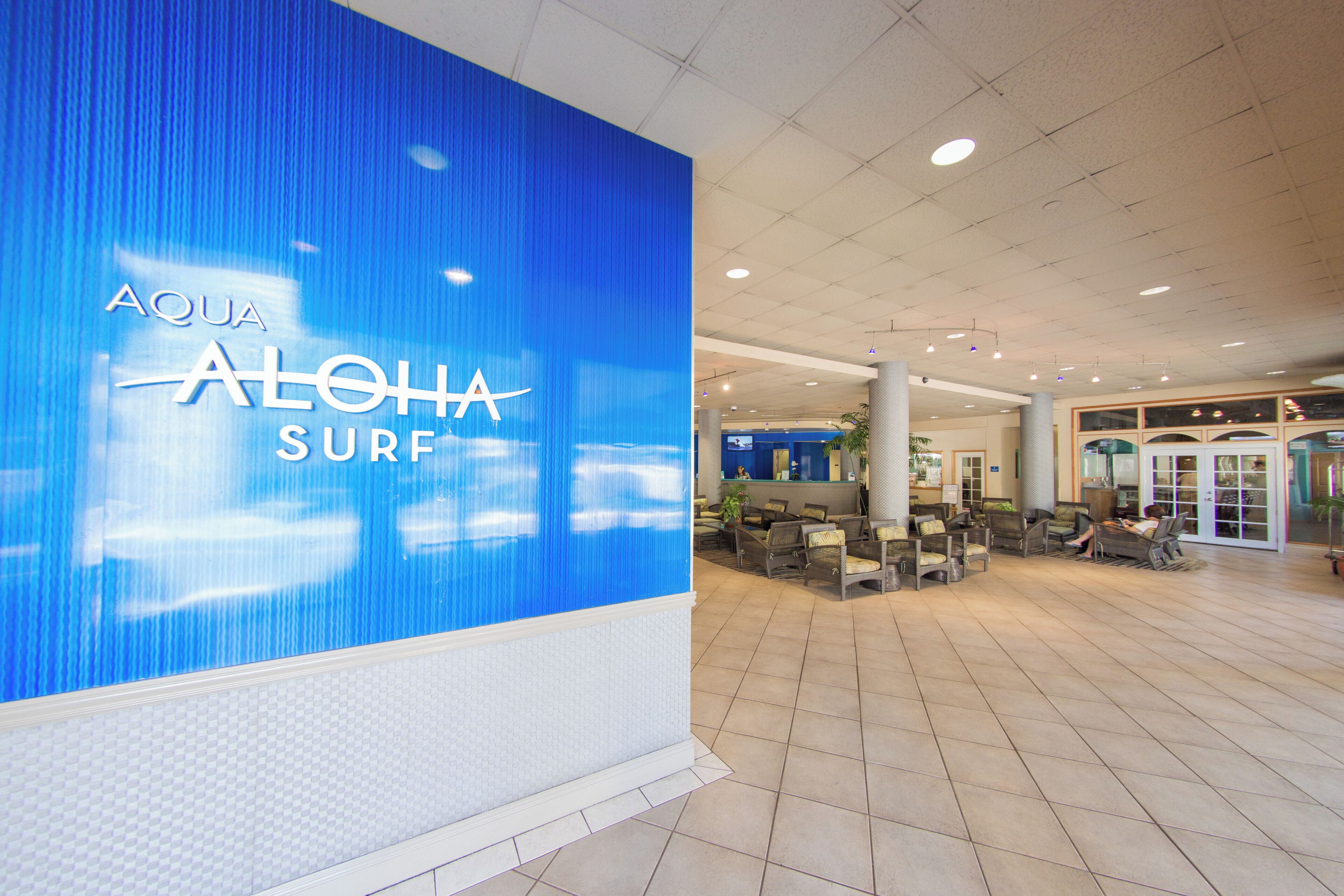 Un vestíbulo amplio con una gran pared azul que muestra 'AQUA ALOHA SURF'. Hay zonas de estar con sillas y mesas, y un mostrador de recepción al fondo.