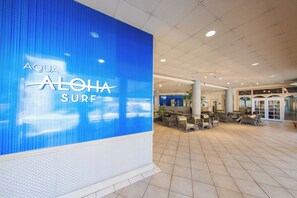 Un hall d’entrée spacieux, avec un grand mur bleu sur lequel est affiché le texte « AQUA ALOHA SURF ». On y trouve des coins salon avec des chaises et des tables, et un comptoir d’accueil en arrière-plan.