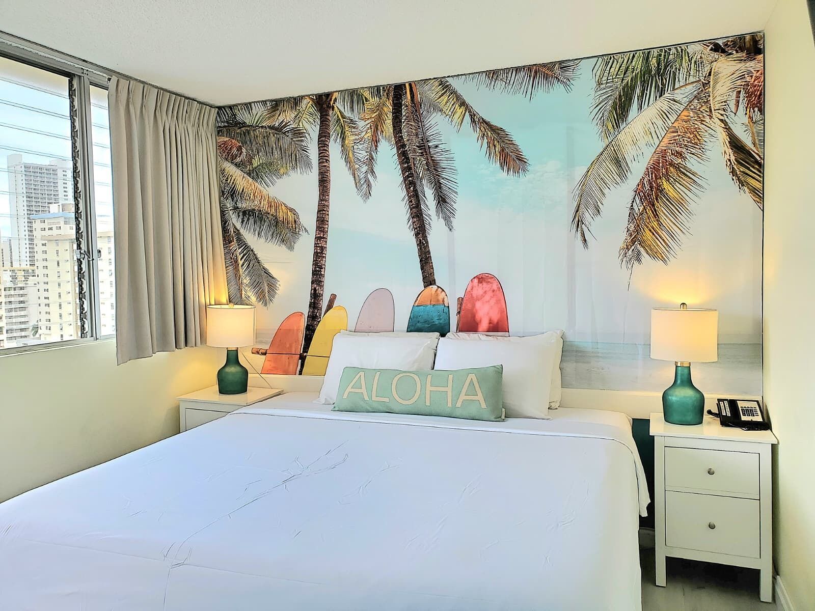 Une chambre d’hôtel avec un lit, deux tables de chevet, une fenêtre avec des rideaux, une vue sur la ville et une fresque murale représentant des palmiers.