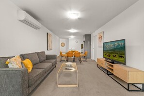 A modern living room with a grey sofa, a glass coffee table, a dining area with orange chairs, a TV on a wooden stand, and an air conditioning unit on the wall. - Exclusive Stays - Mainpoint (Southbank)