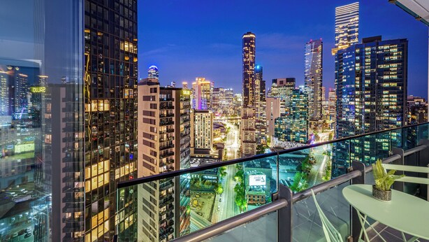 Vista panorámica de la ciudad desde un edificio alto, con varios rascacielos iluminados, barandilla de vidrio y una pequeña mesa con una planta.