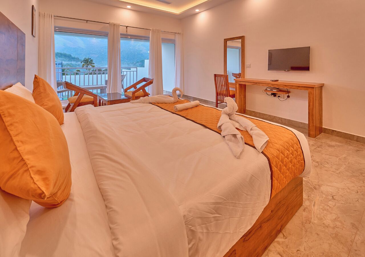A hotel room with a large bed, a desk, a chair, a TV, and a balcony with a view.