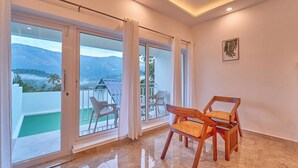 Amplia habitación con un gran ventanal corredero que da a un balcón con vistas a montañas y vegetación. Hay dos sillas de madera y una mesita.