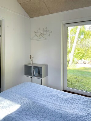 Un dormitorio con cama, una mesita de noche, una ventana con vistas a follaje y un perchero en la pared.