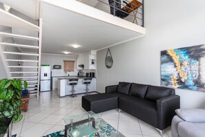 Ein moderner Wohnraum mit einem schwarzen Ecksofa, einem Glastisch, einer Küchenzeile und einer Treppe, die in ein Obergeschoss führt.