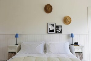 Ein Bett mit weißer Bettwäsche, zwei Nachttische mit Lampen, eine Wand mit zwei Hüten und zwei gerahmten Bildern.