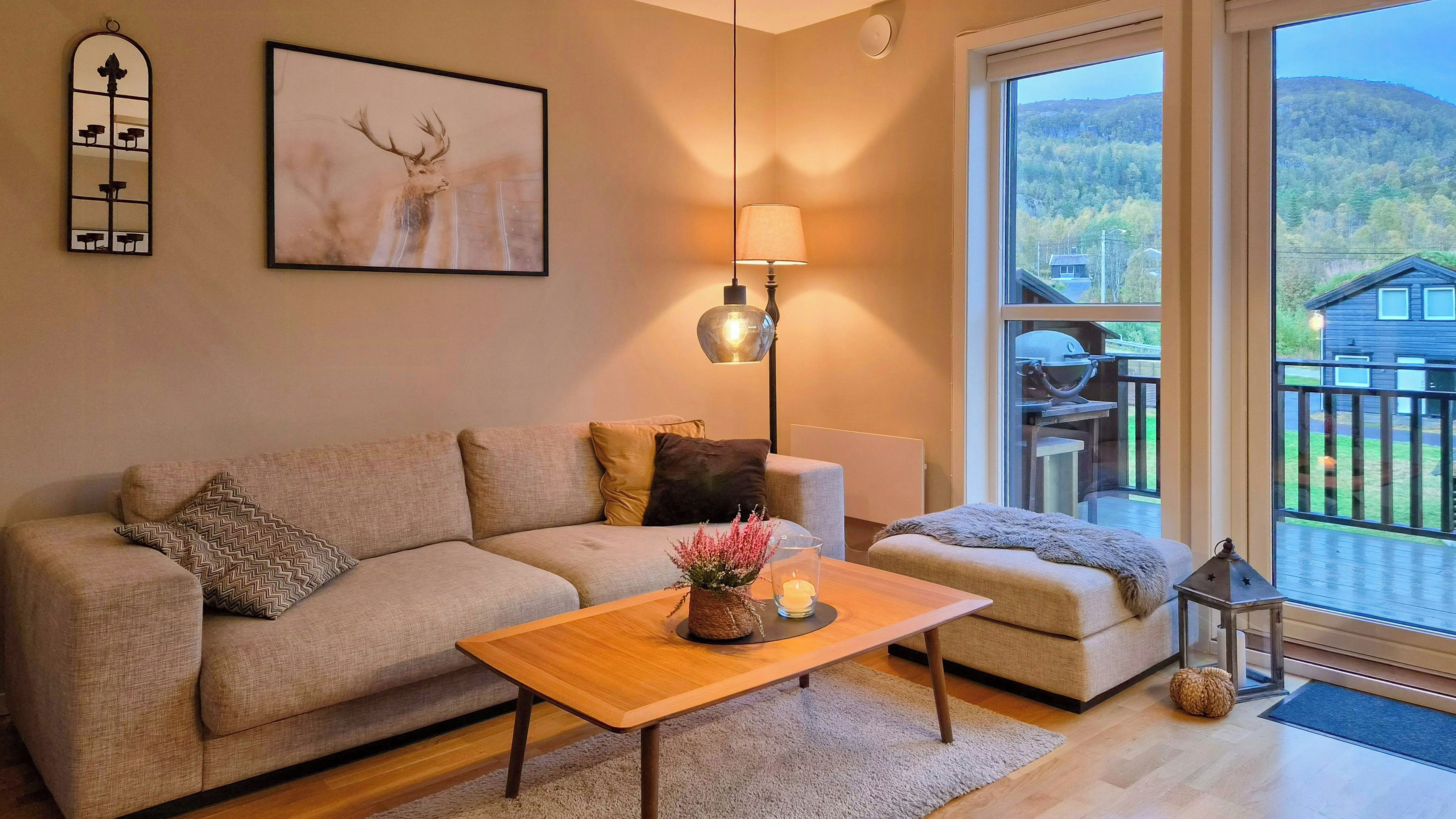 A living room with a sofa, a coffee table, a lamp, and a window with a view of a deck and mountains.