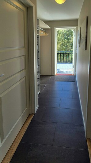 Een gang met een donkere betegelde vloer die leidt naar een deur met uitzicht op een balkon en groen buiten.