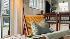 Ein gemütliches Zimmer mit einem Holzstuhl, einem Tisch mit Zeitschriften und Blick ins Freie durch große Fenster.
