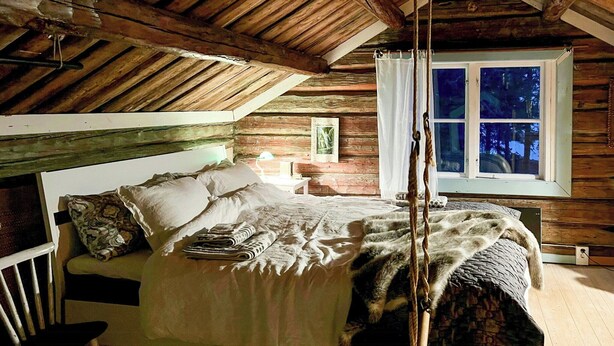 Un dormitorio acogedor en el ático con techo de madera, cama con ropa de cama blanca, un sillón colgante y una ventana con vistas a árboles.