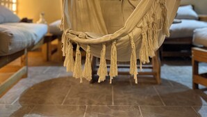 Un'amaca sospesa al soffitto in una stanza con pavimento piastrellato, un letto con biancheria bianca e una finestra con persiane.
