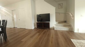 Un ampio soggiorno con pavimento in parquet, una televisione a schermo piatto su un supporto, una zona pranzo con sedie e una scala che conduce a un piano superiore.