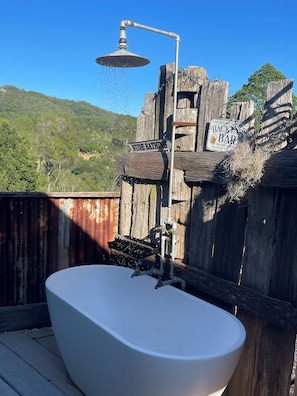 Une baignoire rustique installée à l’extérieur, dotée d’une pomme de douche, entourée de structures en bois et agrémentée d’un panneau indiquant « BAINS NUS ».