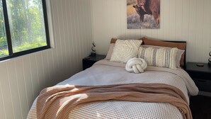 A bedroom with a large window, a bed with a patterned blanket, a wall-mounted picture of a cow, and a nightstand with a lamp.