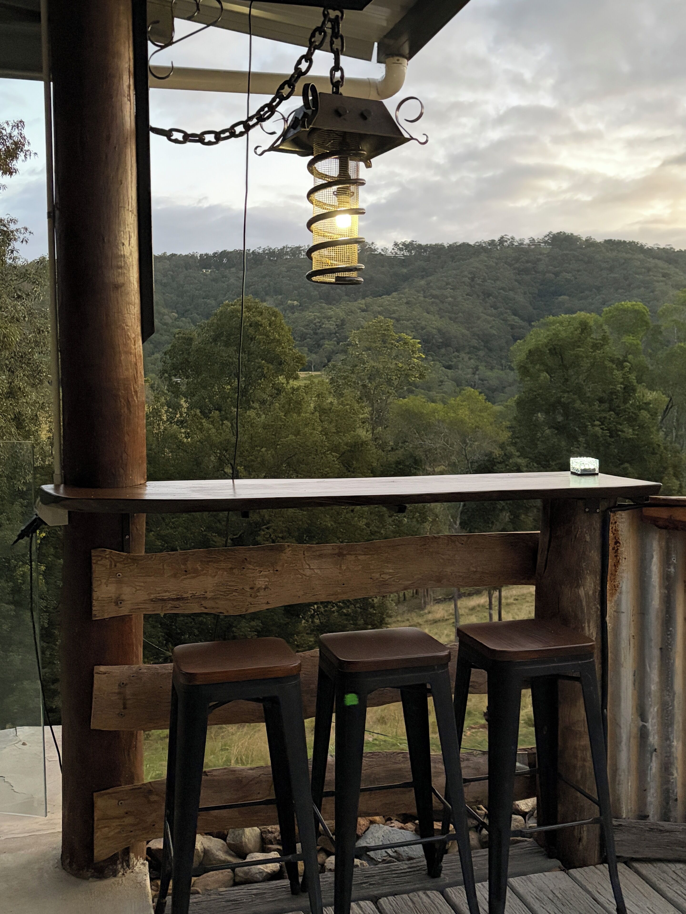 A wooden deck with a metal lantern, two stools, and a view of a forested hillside.