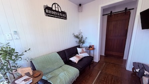 A cozy living room with a black sofa, a green throw blanket, a small table with a book, and a sign on the wall.