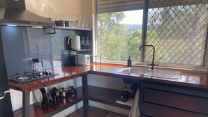 A kitchen with a gas stove, a sink, a window with a view of trees, and a shelf with kitchenware.