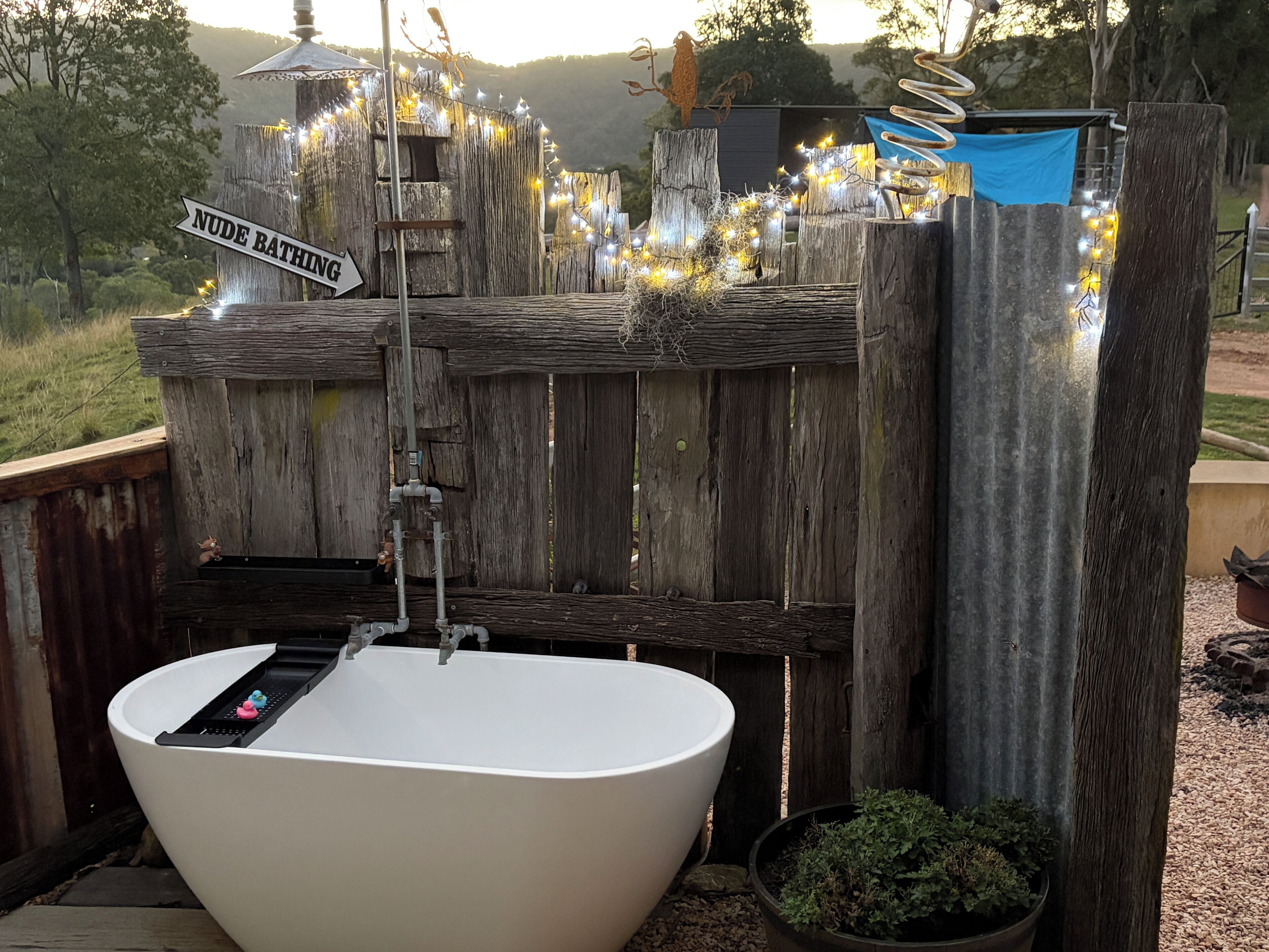 Una bañera rústica al aire libre con un fondo de madera, un letrero que indica 'NUDE BATHING' y luces decorativas.
